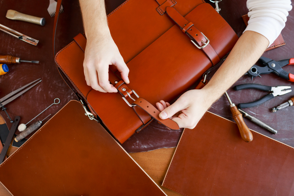 Optimisation des achats et approvisionnements en maroquinerie – artisan ajustant une sacoche en cuir dans un atelier équipé d'outils spécialisés.